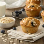 Freshly baked oatmeal muffins on a wooden table with oats and blueberries.