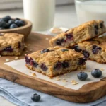A close-up of three golden-brown blueberry oatmeal breakfast bars studded with plump blueberries, arranged on a wooden board with fresh berries scattered around.