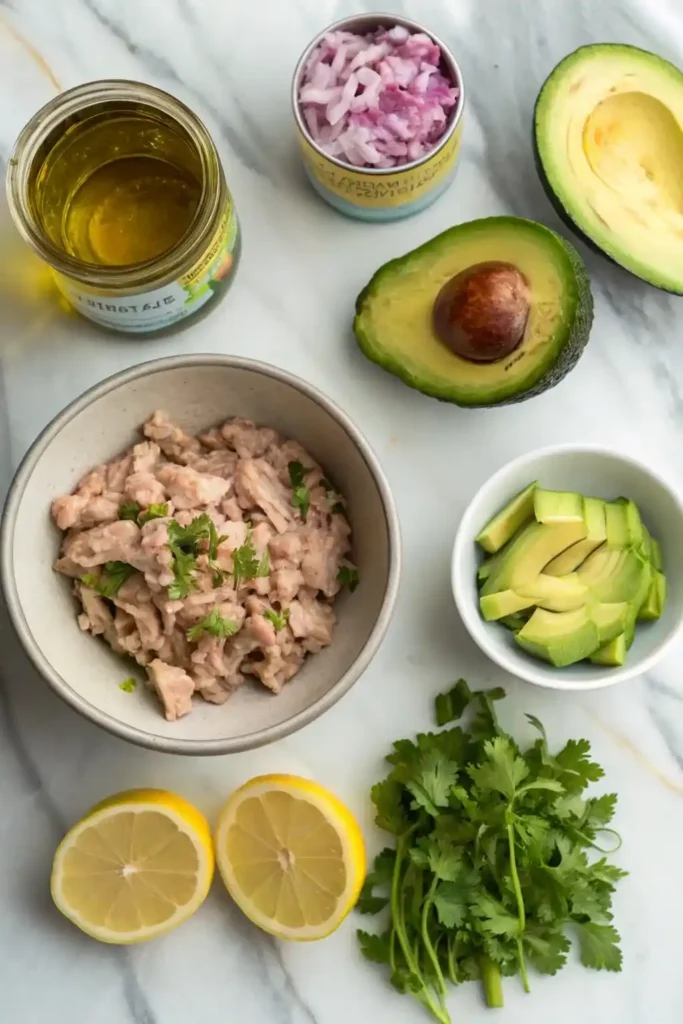 Overhead view of avocado, canned tuna, celery, red onion, lemon, cilantro, and olive oil on a marble countertop.
