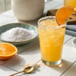 A glass of cortisol cocktail made with orange juice and coconut water on a white counter, surrounded by fresh ingredients and natural light