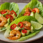 Chili lime shrimp lettuce cups with avocado cubes red chili slices cilantro and lime wedges on a rustic plate.