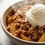 A close-up of a freshly baked healthy apple crisp in a rustic ceramic bowl, topped with a melting scoop of vegan vanilla ice cream. The golden-brown oat and pecan topping is crisp, revealing tender cinnamon apples beneath, all on a distressed wooden surface.