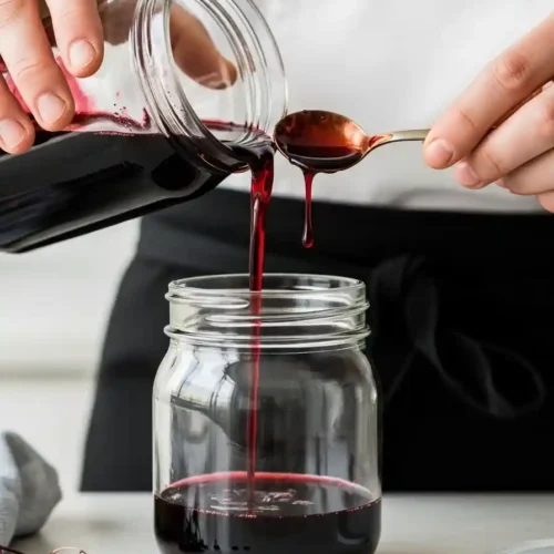 Close-up of a chef carefully pouring homemade elderberry syrup into a clean, airtight glass jar for storage.