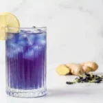 Elegant glass of vibrant blue-purple ACV tonic with lemon garnish on a white marble surface, ready for consumption.