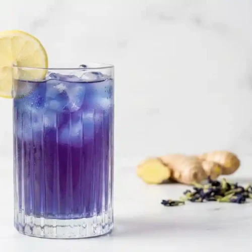 Elegant glass of vibrant blue-purple ACV tonic with lemon garnish on a white marble surface, ready for consumption.