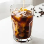 Tall glass of iced cold brew coffee with a visible swirl of oat milk being poured into it, set on a bright white counter.