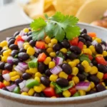 Close-up of vibrant black bean and corn relish in a rustic bowl, garnished with cilantro, with blurred tortilla chips and a taco in the background.Brighten your table with this fresh and flavorful Black Bean and Corn Relish – perfect for any occasion!