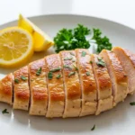 Close-up of perfectly seared thin-sliced chicken breast with fresh lemon wedges and parsley on a white plate.