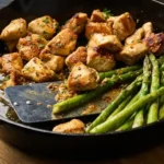A cast iron skillet filled with golden brown chicken bites and bright green asparagus spears coated in a glossy garlic butter sauce, garnished with parsley.
