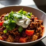 Close-up of a steaming bowl of hearty ground venison chili, garnished with fresh cilantro and sour cream on a rustic wooden table.