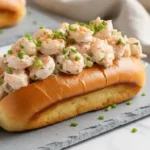 A close-up of a perfectly golden, butter-toasted New England style bun generously stuffed with creamy, herb-flecked langostino lobster meat, garnished with chives and paprika.