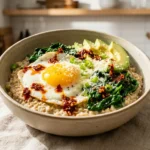 A close-up of a rustic bowl of savory oatmeal topped with a fried egg, sautéed spinach, avocado, Parmesan, scallions, and chili crisp on a linen tablecloth.