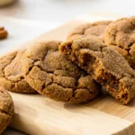 Close-up of three soft and chewy molasses cookies, one broken open, on a wooden board, showing crinkled tops and chewy interior.