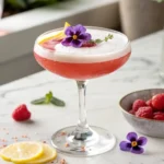 A beautifully garnished Raspberry Passionfruit Tequila Sour in a coupe glass on a light background, featuring a lemon twist, fresh raspberry, and edible flower.