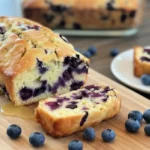 A golden-brown, sliced blueberry cream cheese loaf on a rustic wooden cutting board, revealing a moist interior with blueberries and cream cheese swirls.