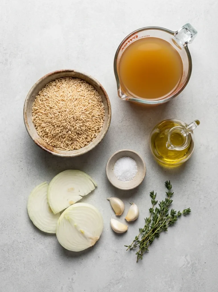 Flat-lay arrangement of raw brown rice, vegetable broth, olive oil, salt, onions, garlic, and thyme on a light background.