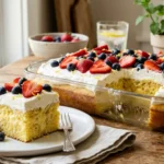 A close-up of a slice of lemon berry poke cake with whipped cream, strawberries, and blueberries on a white plate.