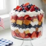 A tall glass trifle dish showing patriotic layers of red strawberries, white cream, and blue blueberries.Dessert