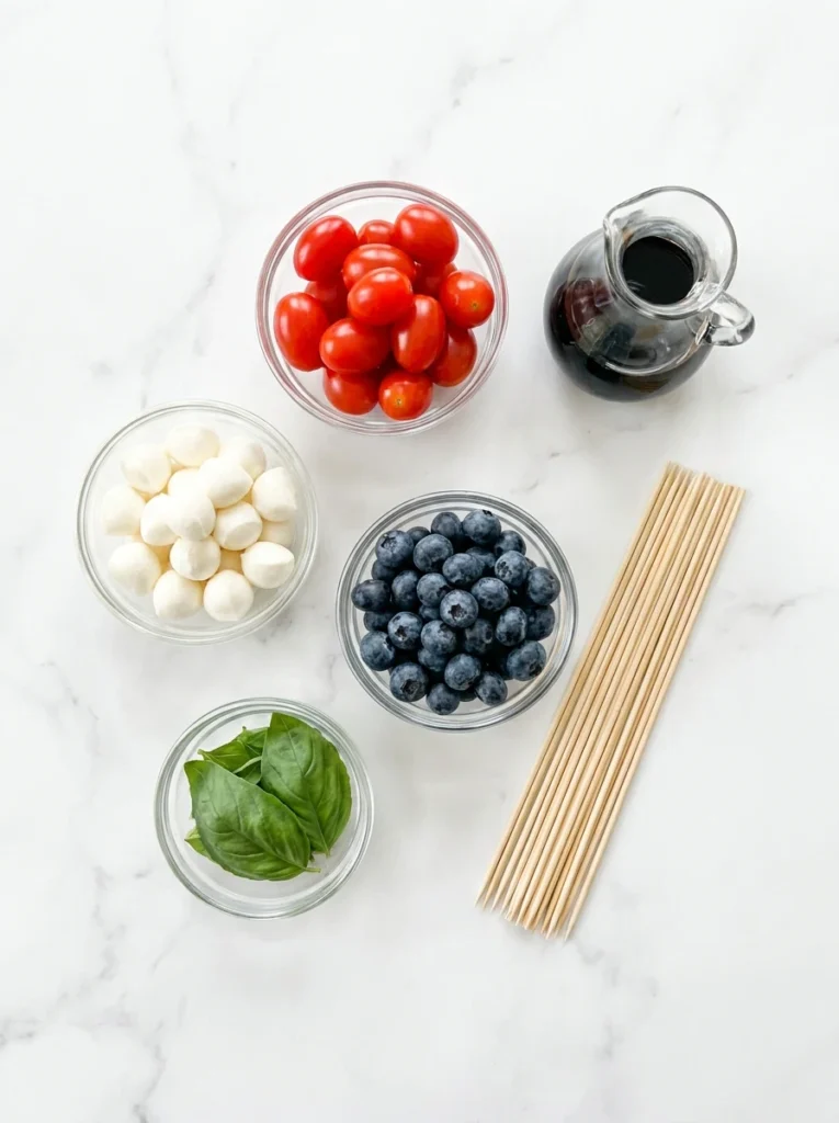 Individual bowls of cherry tomatoes, mozzarella pearls, blueberries, and basil leaves on a white marble background.