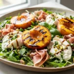 A close-up overhead shot of a vibrant salad featuring roasted peaches, creamy burrata cheese, prosciutto, and arugula with balsamic glaze.