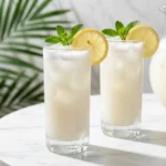 Two glasses of frothy white coconut lemonade with mint and lemon garnishes on a marble table.
