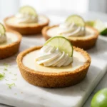 A group of mini key lime pies topped with whipped cream and lime slices on a white marble surface.