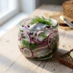 A close-up of a glass jar filled with pickled herring, red onions, and fresh dill on a wooden table.