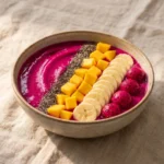 A beautifully styled magenta-colored dragon fruit smoothie bowl topped with chia seeds, mango, and sliced bananas.