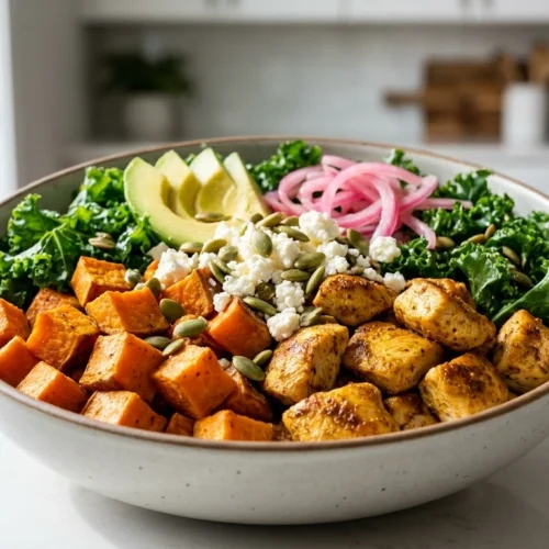 Sweet Potato And Chicken Bowls .A colorful grain bowl with roasted chicken, sweet potatoes, kale, avocado, and pickled onions on a white ceramic plate.