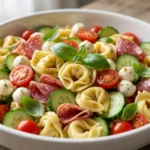 A large bowl of colorful tortellini pasta salad with tomatoes, mozzarella, and salami on a rustic wooden table.