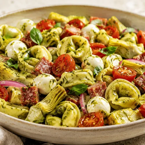 A large bowl of pesto tortellini salad with mozzarella pearls, tomatoes, artichoke hearts, and beef salami on a stone countertop.