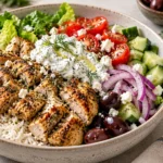 A professionally styled Greek chicken bowl with seared chicken, basmati rice, romaine lettuce, fresh vegetables, feta, and tzatziki sauce on a stone countertop.