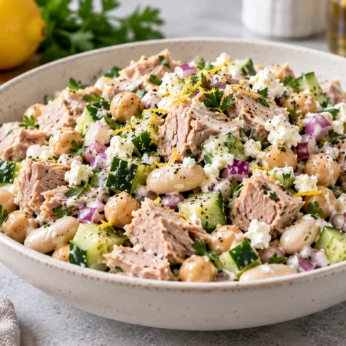 A vibrant, high protein tuna salad with chickpeas, cannellini beans, cucumbers, and feta cheese in a white bowl on a stone countertop.