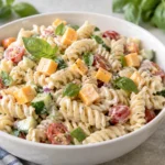 A large serving of kid-friendly pasta salad in a white bowl, garnished with fresh basil leaves and cubed cheddar cheese