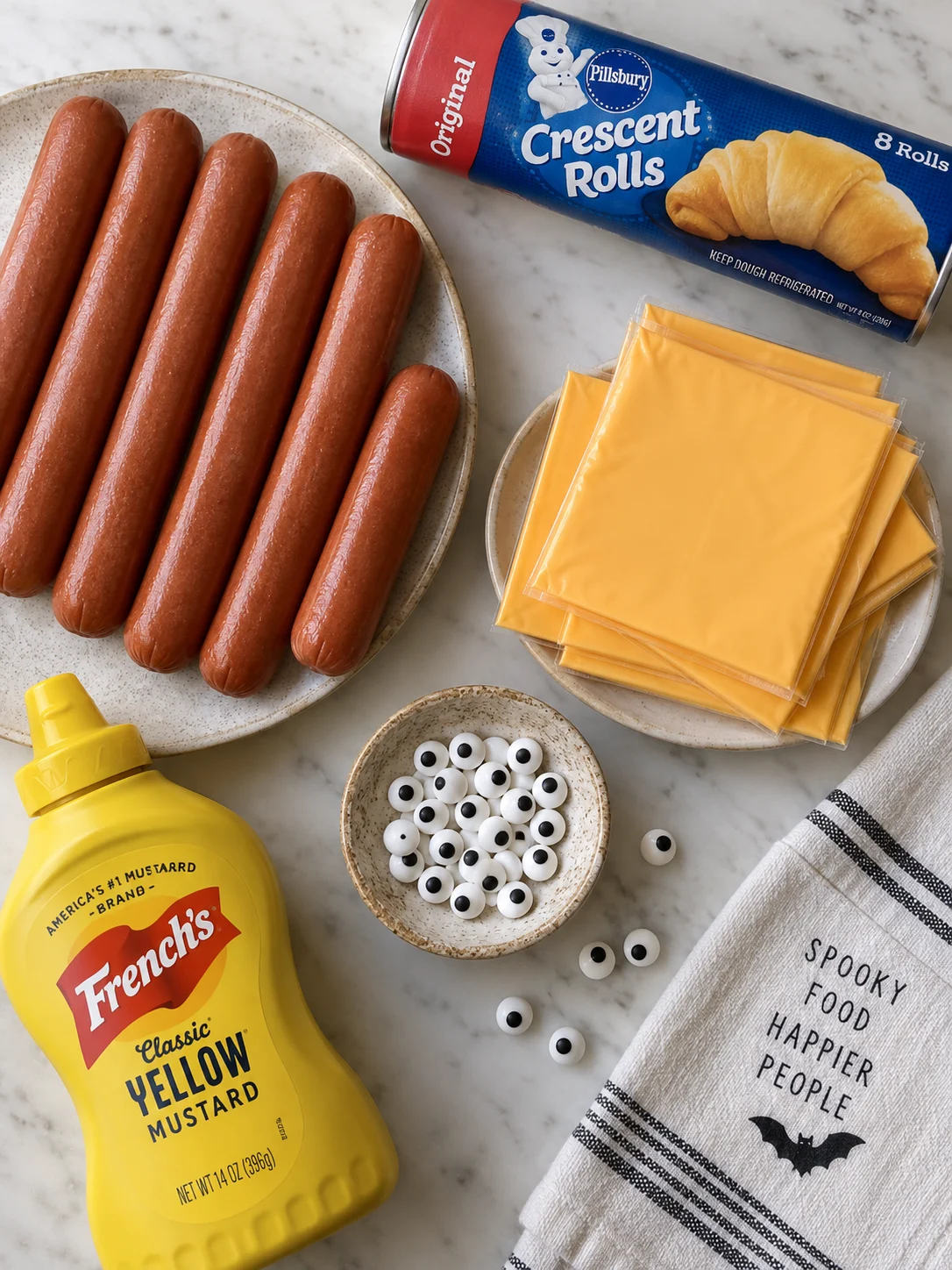 A flat-lay of ingredients for mummy hot dogs, including beef hot dogs, crescent roll dough, and candy eyes