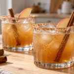 Two glasses of amber-colored apple cider mocktail with cinnamon-sugar rims and apple slice garnishes on a wooden table.