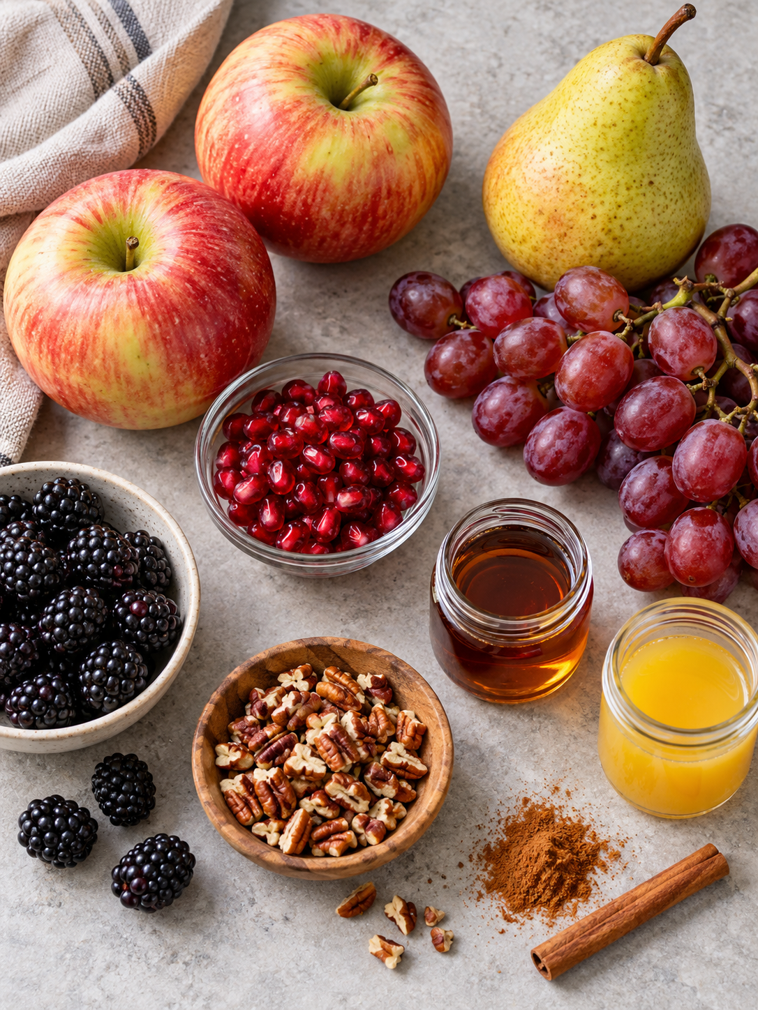 Top-down view of whole apples, pears, grapes, and bowls of pecans and pomegranate seeds arranged on a stone surface.