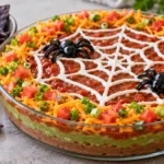 A layered taco dip in a glass dish topped with a sour cream spider web and olive spiders, surrounded by blue corn chips.